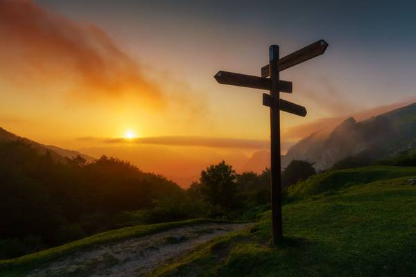 Sonnenaufgang oder -untergang mit leichtem Nebel in den Bergen. Auf einer Wiese steht ein Wegweiser, der in verschiedene Richtungen zeigt.