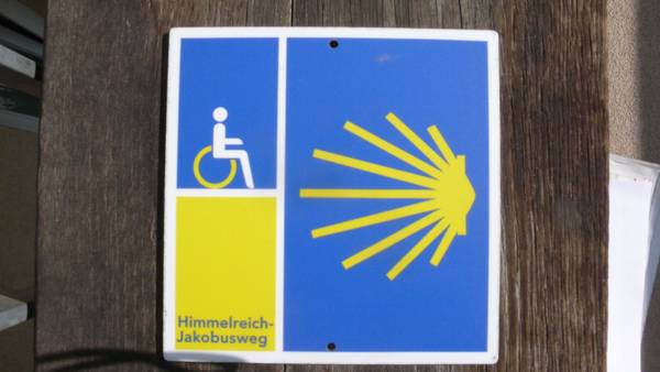 An einem Pfosten befindet sich ein Schild mit der Jakobusmuschel der Pilger*innen, einem Rollstuhlsymbol und dem Schriftzug Himmelreich-Jakobusweg.