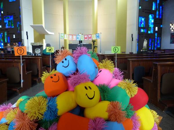 Es liegen bunte Knautschbälle mit Gesichtern auf einem Haufen in einer Kirche. Die Bankreihen sind für Menschen mit unterschiedlichen Behinderungen ausgeschildert.