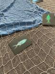 Auf dem Foto ist ein Fischernetz zu sehen. Dieses liegt teilweise auf einem blauen Tuch und teilweise auf dem Teppichboden. Auf dem Netz liegen wiederum zwei Postkarten, auf denen jeweils ein gemalter Fisch auf schwarzem Hintergrund abgebildet ist.