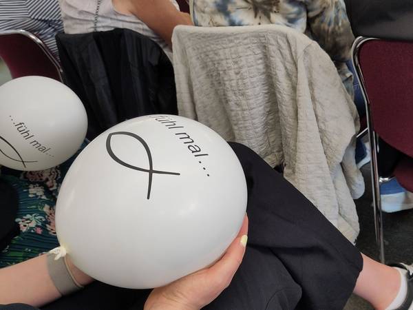Ein weißer Luftballon wird auf dem Schoß in der Hand gehalten. Darauf steht geschrieben: Fühl mal... darunter ist das Piktogramm eines Fisches. Im Hintergrund ist der selbe Luftballon und die Rückenlehnen der vorderen Stuhlreihe zu sehen.