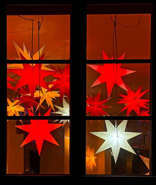 Bunte Sternleuchten hinter einem Sprossenfenster im Innern eines Hauses.